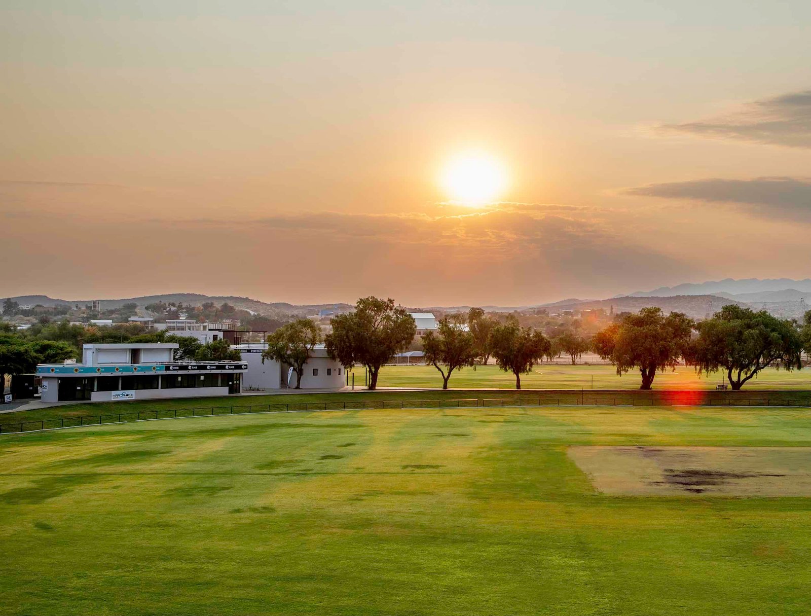 Namibia Cricket Ground in Windhoek at sunset, with the outfield, pitch, clubhouse and surrounding hills.