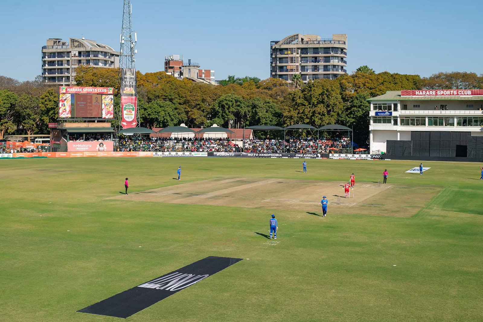 Harare Sports Club during a cricket match, showing the central pitch, pavilion, scoreboard and tree-lined boundary.