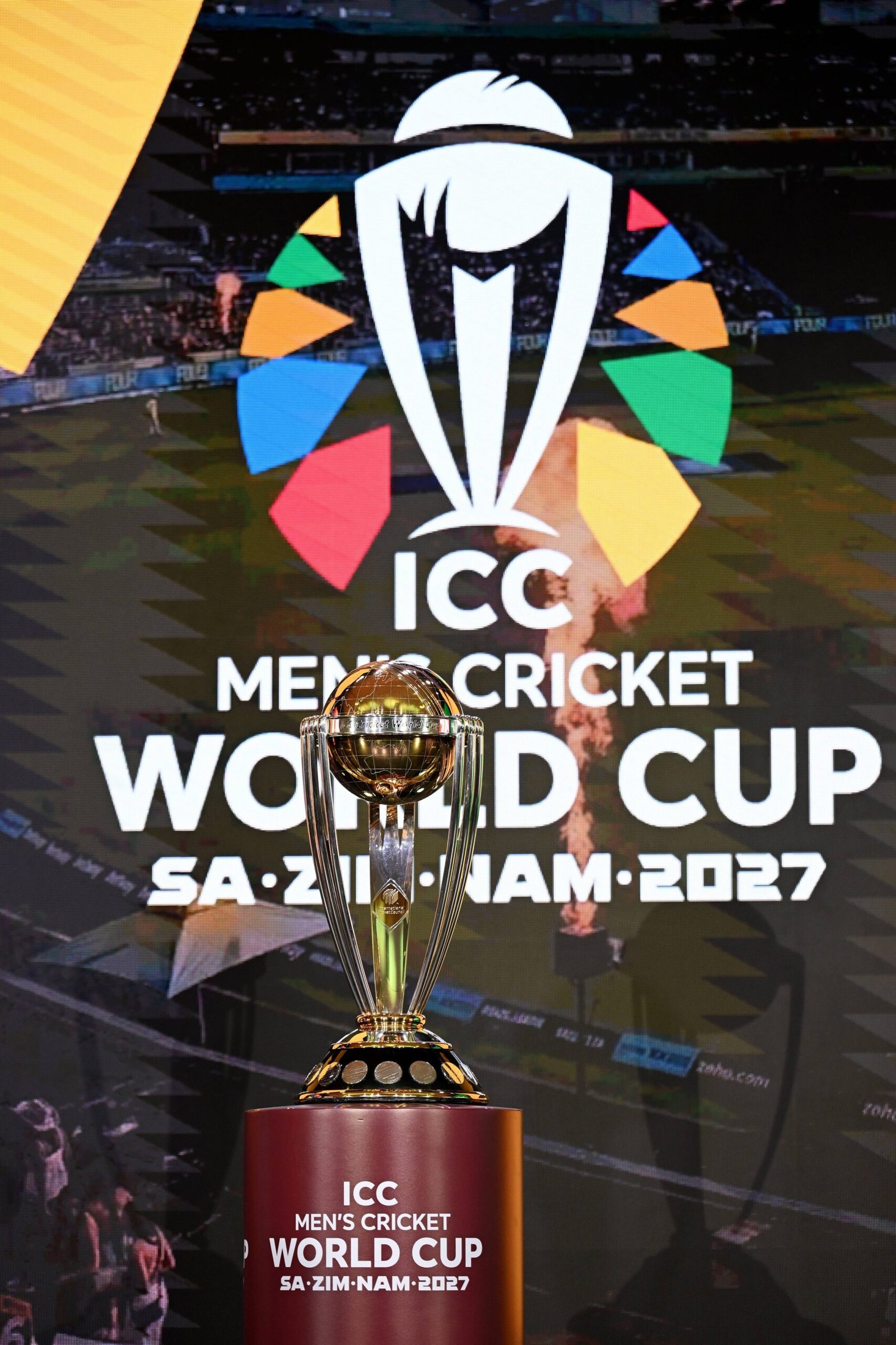 ICC Men’s Cricket World Cup trophy displayed on a podium against the 2027 tournament branding for the guide to host stadiums in South Africa, Zimbabwe and Namibia.